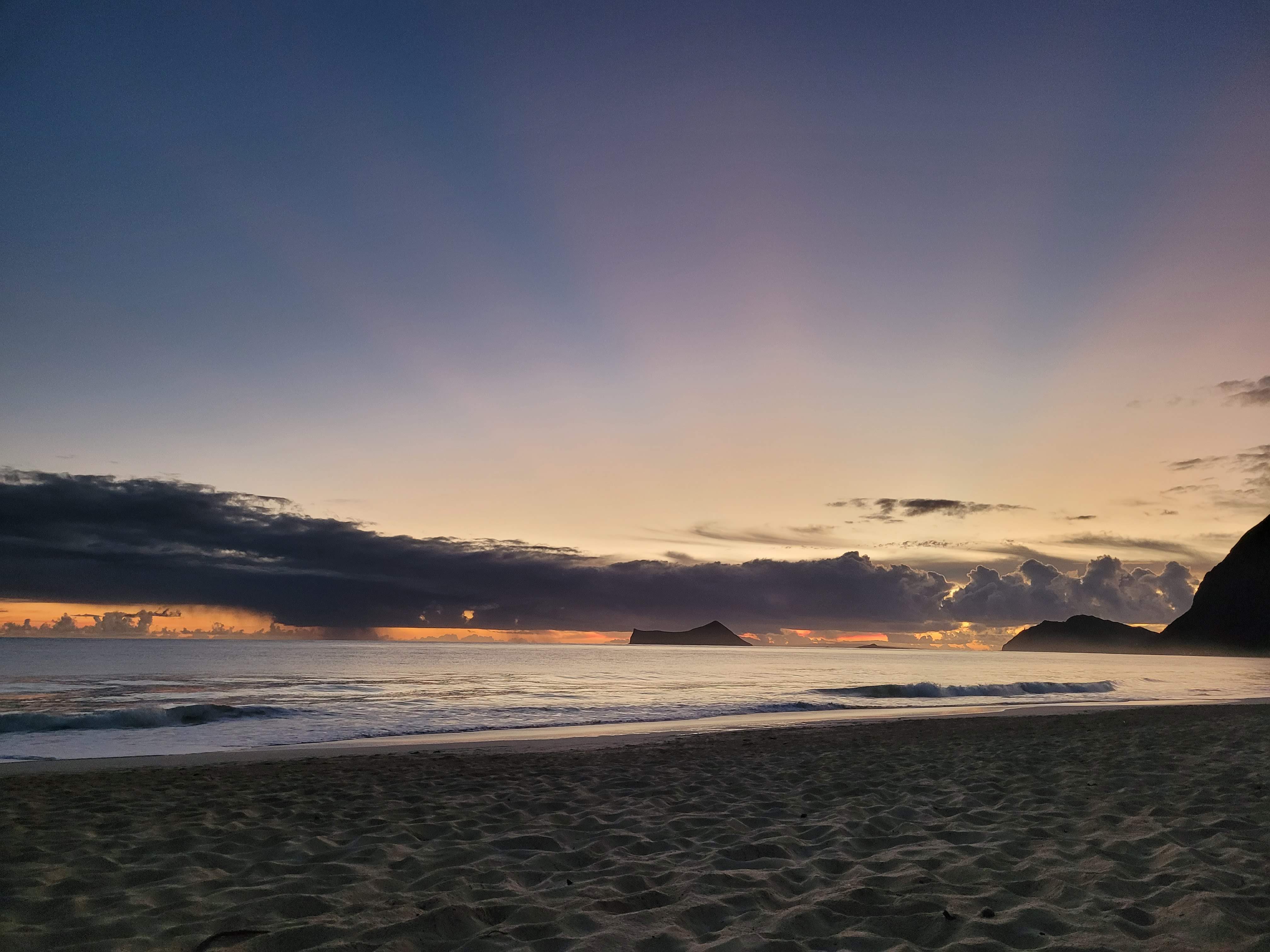 A gentle sunset over a calm ocean, with a sandy beach foreground and the silhouette of a distant island.
