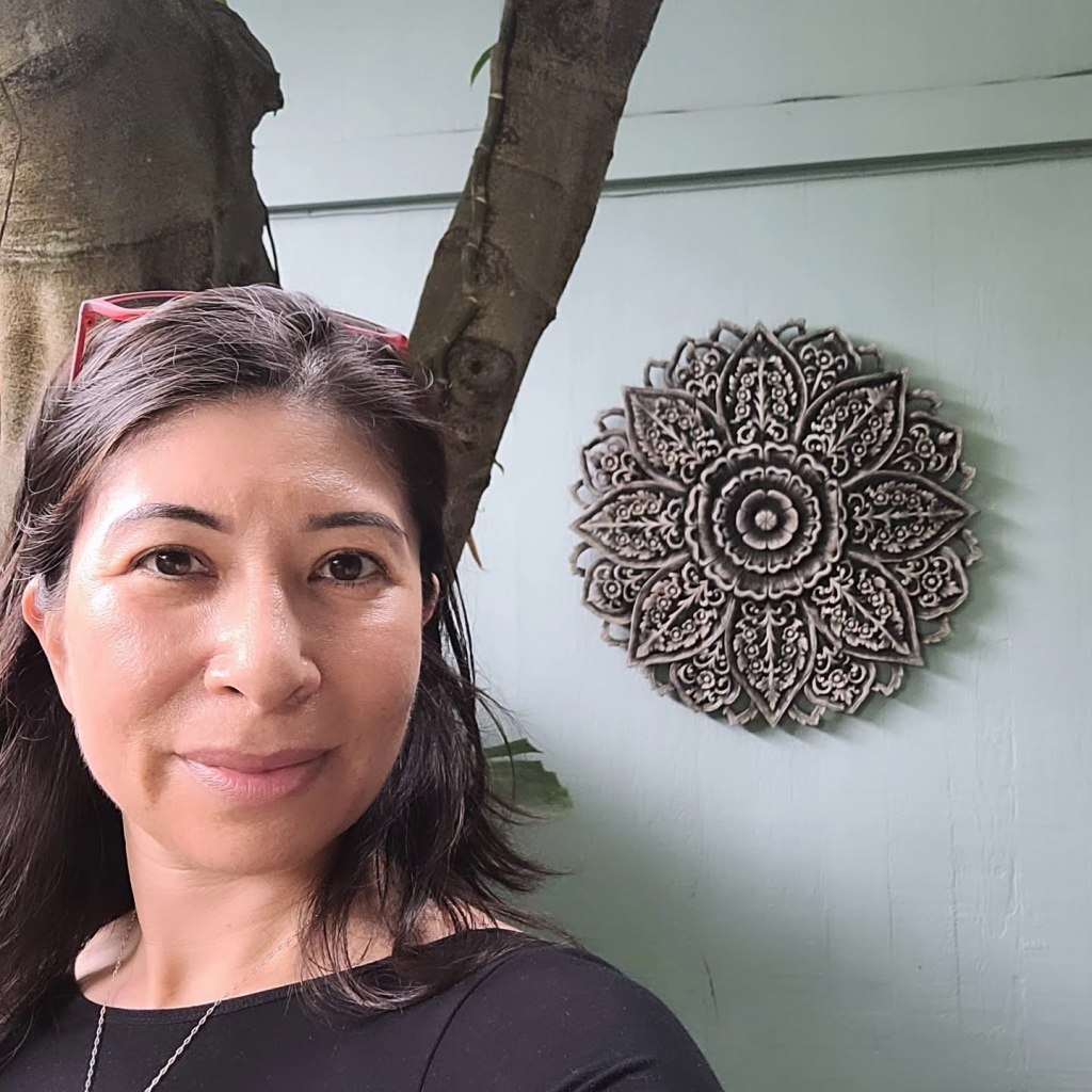 A woman with medium-length, dark hair and a black shirt stands in front of a light green wall, adorned with a gray mandala.